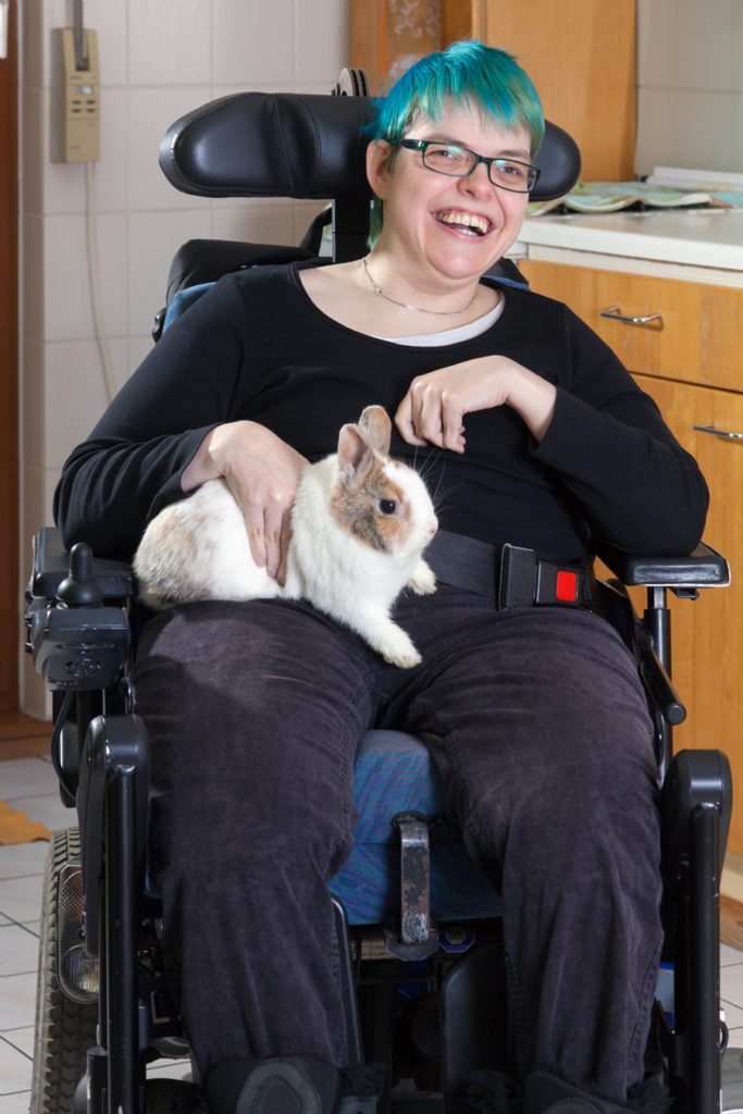 Cheerful young infantile cerebral palsy patient caused by complications at birth sitting in a multifunctional wheelchair stroking a pygmy rabbit as therapy, with a beaming smile.